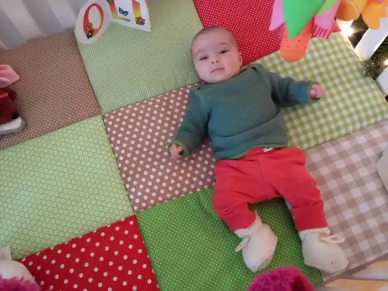Een baby die een groen shirt, rode broek en witte sloffen draagt, ligt op een gekleurd gestikt dekbed.